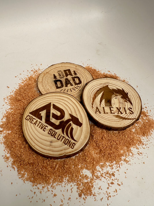 Three pine coasters with words or images laser engraved laying on a bed of sawdust
