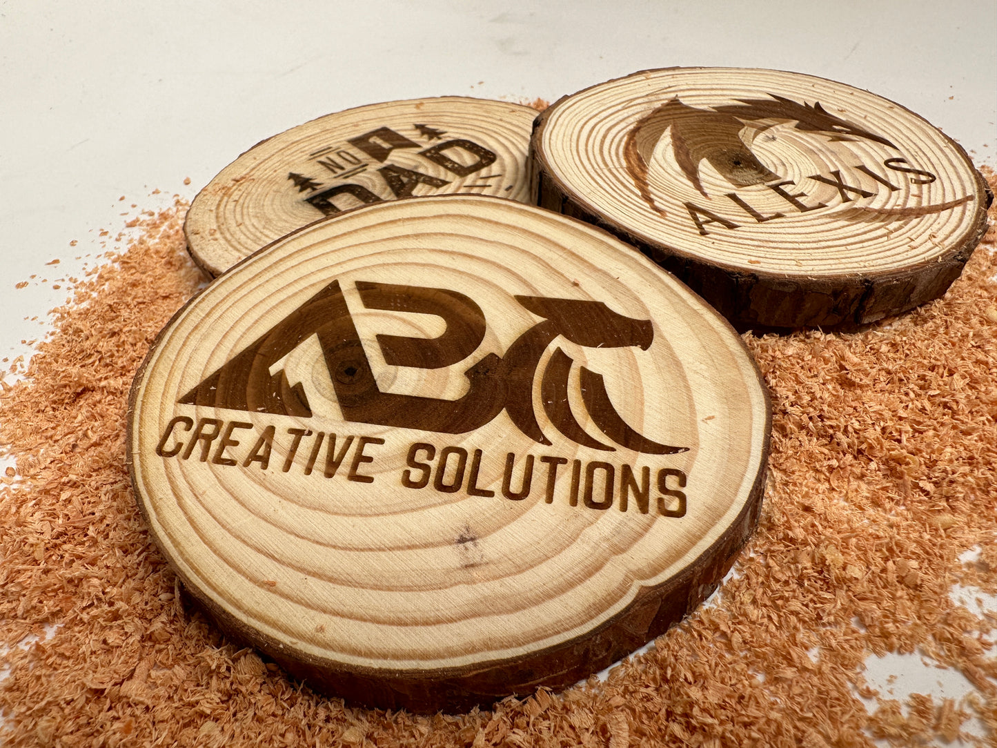 A close up view of three wooden coasters laser engraved with words or images on a pile of sawdust